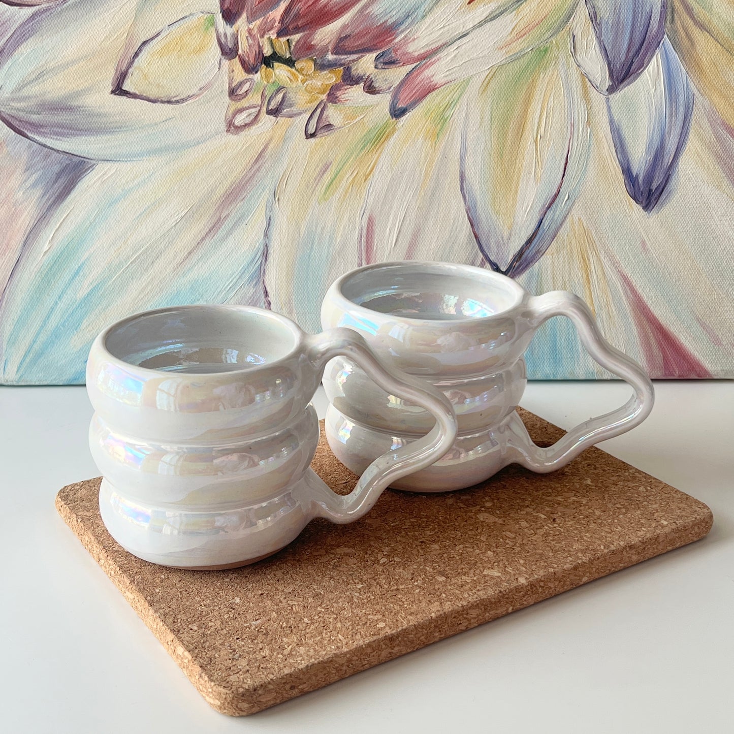 Two handmade ceramic mugs with iridescent white glaze and wavy handles, displayed side by side on a corkboard.