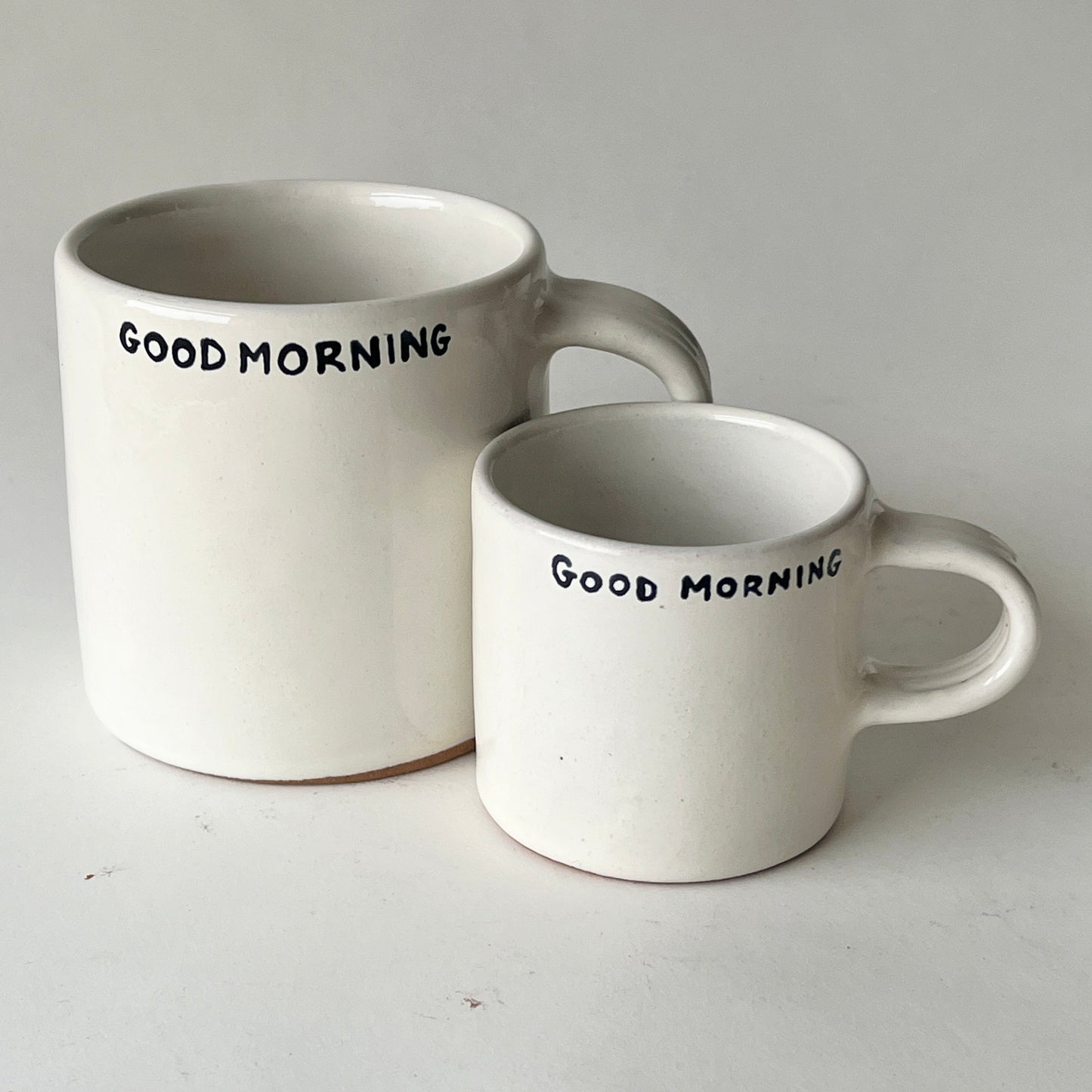 A pair of handcrafted ceramic mugs in two sizes, both with a simple "GOOD MORNING" message in black, perfect for gifting.