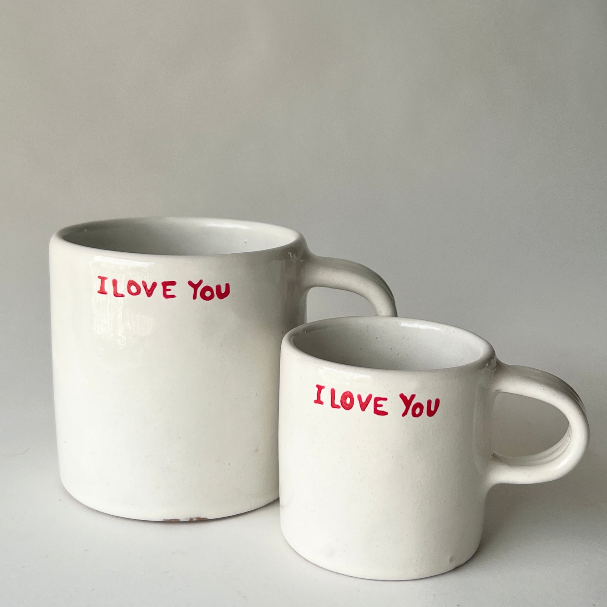 Two handmade ceramic mugs with an off-white glaze, featuring "I LOVE YOU" hand-printed in red, displayed side by side.