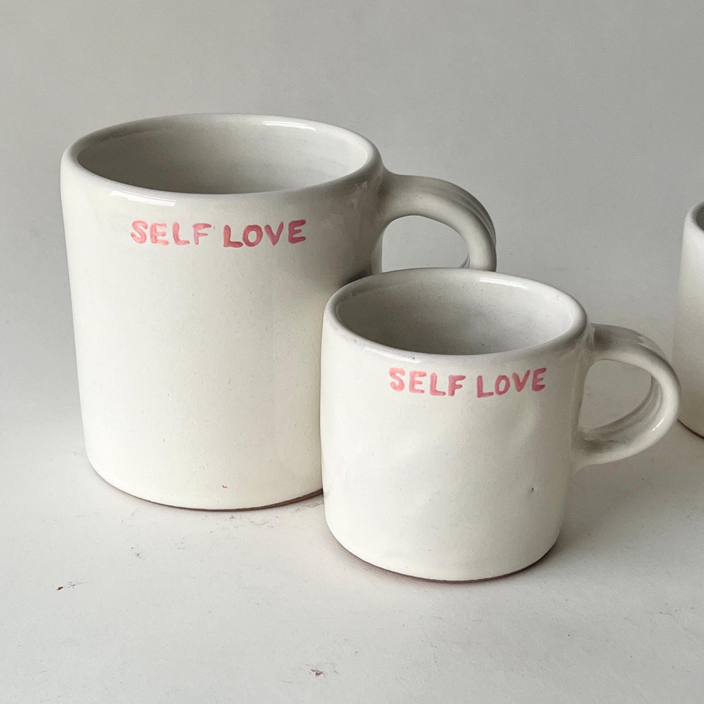 Two handmade ceramic mugs with an off-white glaze, featuring "SELF LOVE" hand-printed in pink, displayed side by side.